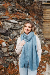Woman wears pale blue crochet scarf, wrapped with ends hanging, in front of stone wall. thumbnail