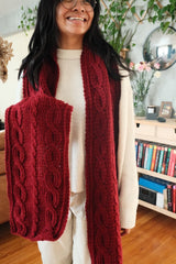 Smiling woman holds end of cranberry red crochet scarf in hand, with other end hanging free. thumbnail