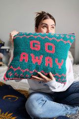Woman on bed holds a green crochet pillow. Text in pink reads Go Away, and there is a border at the top and bottom. thumbnail