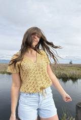 Woman stands in nature preserve, with wind blowing her hair in her face. She has a crochet lace top on, with a drawstring bottom, and pale denim shorts. thumbnail