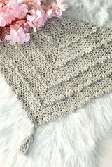 Flat lay of romantic lace crochet shawl, with tiered scalloped ruffles and a tassel at the center point. It's laid on a white faux fur rug, with a bouquet of pale pink flowers in the upper left corner. thumbnail