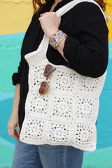 Close up of crochet shoulder bag, made of individual squares of cream yarn. Heart-shaped aviator sunglasses hang from the top of the bag. thumbnail