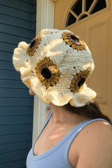 Back view of crochet sun hat showing sunflower granny square on top, and around the body of the hat. The flowers have a black center, then brown, then mustard yellow. The space between flowers is cream, and the brim is ruffled. thumbnail