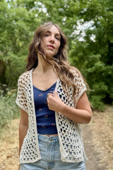Woman in a blue top and jeans, with wind blowing her hair, standing on a dirt path through trees. She also wears a cream crochet sleeveless cardigan. thumbnail