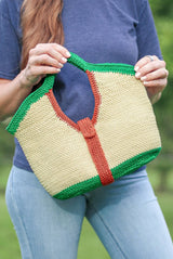 Woman holds crochet purse at jaunty angle. The purse has bright green trim at top and bottom, red-orange trim around the inner handle and as a vertical strap around the middle. The body of the bag is a straw yellow color. thumbnail
