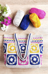 Top down view of crochet tote bag on wooden table top. The bag handles are folded down, and there are flowers and yarn skeins above the bag on the table top. thumbnail