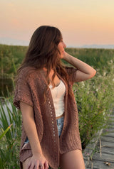 Front side view of woman sitting on the edge of a boardwalk in a wetland area, looking over her left shoulder. She wears an open short sleeve cardigan over a tank top and jean shorts. thumbnail