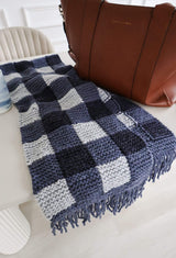 Gingham knit scarf laid on counter with a brown shoulder bag in the background. thumbnail