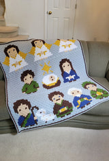 Afghan craped over grey couch indoors. The afghan has a pale blue background with three rows of cartoon style figures from the nativity scene. thumbnail