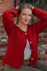 Woman sitting on brick steps wearing red-orange cardigan. She has her arms raised and the cardigan its buttoned at the very top. thumbnail
