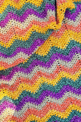 Top down view of crochet chevron blanket in self-striping yarn. The yarn is mustard yellow, green, purple, beige and orange. thumbnail
