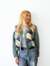 Front view of woman standing in front of a white brick wall. She wears a crochet cardigan in blue-green, dark denim, and cream. The sleeves are solid and the back has a 3-dimensional design made of granny motifs.  thumbnail