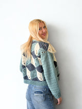 Back view of woman standing in front of a white brick wall. She wears a crochet cardigan in blue-green, dark denim, and cream. The sleeves are solid and the back has a 3-dimensional design made of granny motifs.  thumbnail