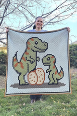 Woman holds up crochet afghan with t-rex family while standing outdoors. thumbnail