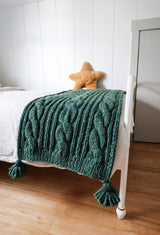 Green cabled throw on a white bed, with a golden star pillow in background. thumbnail