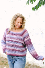 Woman with blonde curly hair wears pink & purple crochet sweater with blue jeans. thumbnail
