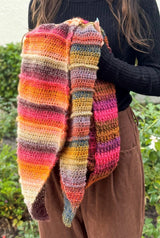 Woman wearing black turtleneck and brown pants holds multicolor crochet shawl in front of her. thumbnail