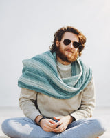 Bearded man in sunglasses, beige sweater, pale blue pants, sits cross-legged. He wears a crochet wrap in shades of blue-green.  thumbnail