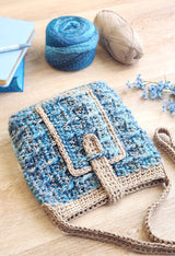 Textured crochet bag laying on wooden table, opening facing the camera. Yarn balls and a stock of journals in the background.  thumbnail