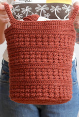 Woman holds tall crochet basket toward camera, holding it by two side handles.  thumbnail