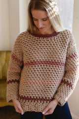 Woman wearing tan crochet sweater with pink stripes. She stands inside with a green couch in background  thumbnail