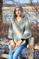 Woman sits on wooden fence in front of snowy field, wearing crochet poncho with hood worn up.  thumbnail