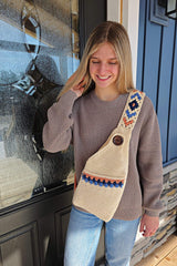 Woman in a tan sweater and jeans stands in front of a house door. She wears a crocheted sling bag in cream, with a pocket on the front and a large button. The strap has a geometric design in navy, blue, and orange.  thumbnail