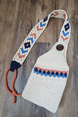 Flat lay of a crochet sling bag. The main bag is cream and has a big button closure. The strap has a geometric pattern in navy, blue, and orange. There is an orange cord to adjust the strap length.  thumbnail