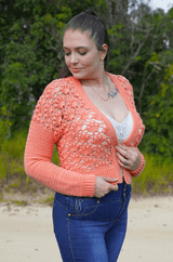 Woman stands on a sandy road, with greenery in the background. She wears a white camisole and blue jeans, and a coral lace crochet cardigan.  thumbnail