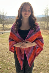 Young woman stands in front of early spring yard, wearing all black except for a bright shawl. The crochet shawl is make in shades of orange, pinks, and purple.  thumbnail