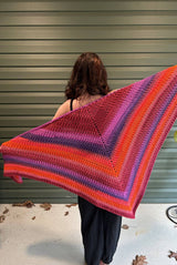 Back view of young woman holding triangular shawl with top edge stretched out. She's standing on a cement porch. The shawl starts with red-purple at the top and stripes through pink, purple, and bright orange.   thumbnail