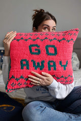 Woman seated on bed holds up crochet pillow. The background is pink and the text in green reads Go Away, with a border top and bottom thumbnail