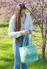 Woman in cream sweater and jeans stands outdoors in front of flowering tree. She holds a green crochet toe bag with long shoulder straps.  thumbnail