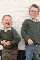 Two young boys wear matching knit sweaters, posed outside against a white painted brick wall. The sweaters are a dark green with saddle shoulders and ribbed collar, cuffs, and hem.  thumbnail
