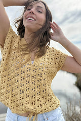 Close up of crochet lace tee, with a cinched bottom hem. The tee is made in a straw yellow color, and the woman wearing it has her arms raised.  thumbnail