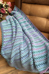 Crochet blanket in pale blue and shades of green and purple, draped over a tan armchair. There is a bouquet of mauve roses in the background.  thumbnail