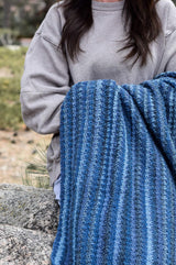 Cropped view of a woman in a grey sweatshirt holding a crochet blanket. She is sitting on a boulder outside, and the blanket is worked in shades of blue.  thumbnail