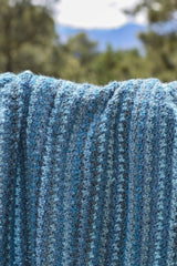 Close up of crochet blanket in shades of blue with trees and a mountain in the distance.  thumbnail