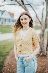 Woman in a pale yellow short sleeve cardigan over a white t-shirt, with pale blue jeans. She stands in a mulches area in front of a bare tree, with a house in the background.  thumbnail