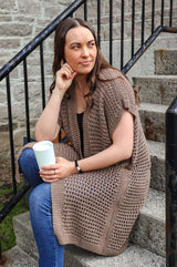 Woman in a long taupe knit cardigan and jeans sits on cement steps, holding a travel mug.  thumbnail