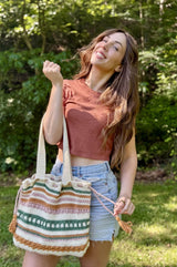 Smiling woman stands in a backyard in front of a treeline. She holds a crochet tote bag in the crook of her elbow. The bag is cream with stripes of amber, sage green, and pale pink.  thumbnail