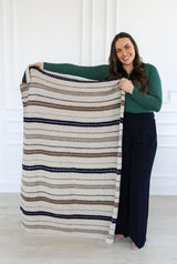 Woman in a green top and black pants stands in front of a white wall. She holds a small throw blanket crocheted in cream, taupe, brown, and blue.  thumbnail