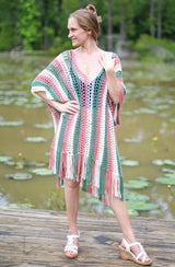 Woman stands on wooden dock above pond with many lily pads. She wears white sandals and a crochet poncho that hangs to mid thigh, with fringe to the knee. The poncho has a v-neck and vertical stripes of cream, bright sage green, and terracotta pink.  thumbnail