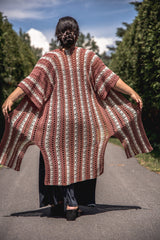 Back view of long crocheted cardigan  worn by woman, with the sides held open. The cardigan has slit sides at the bottom, and vertical stripes of cream, beige, and red-orange.  thumbnail