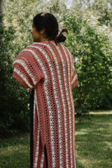 Side view of woman with hand on hip nearest the camera, wearing a long crochet cardigan. You can see the slit at the side hem, and the vertical stripes of lace and color.  thumbnail