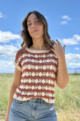 Woman stands in a grassy field under a blue sky. She wears denim shorts and a striped crochet top in cream and russet.  thumbnail