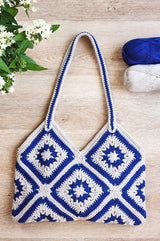 Crochet bag made of many squares seamed together laid on a pale wooden surface, with green and white flowers in the top left and two balls of yarn in the top right. The bag is made in blue and off white and has a striped shoulder length strap.  thumbnail