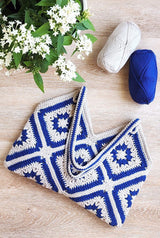 Bag laying on wooden surface with handles folded over bag body. There is a potted plant with green leaves and white flowers in upper left corner, and two balls of yarn in upper right.  thumbnail