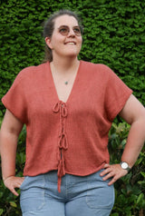 Woman in a red-orange top and striped jeans stands in front of a green hedge. The top has short sleeves and three bows to close the front.  thumbnail