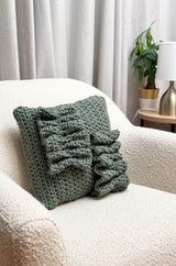 Sage green crochet pillow sits in a ream armchair. The pillow has two ruffle appliques. thumbnail
